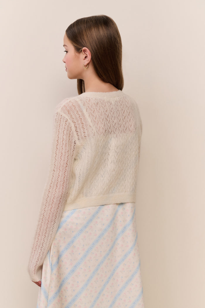 Girl wearing a beige knitted cardigan with ruffles on a plain background
