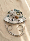 A top-down photograph of a baby bucket hat featuring a light grey or beige fabric covered in a playful zoo animal print, including a zebra, tiger, alligator, and elephant. The hat includes matching fabric chin tie straps and is styled flat on a neutral, textured beige linen background under soft, warm sunlight.