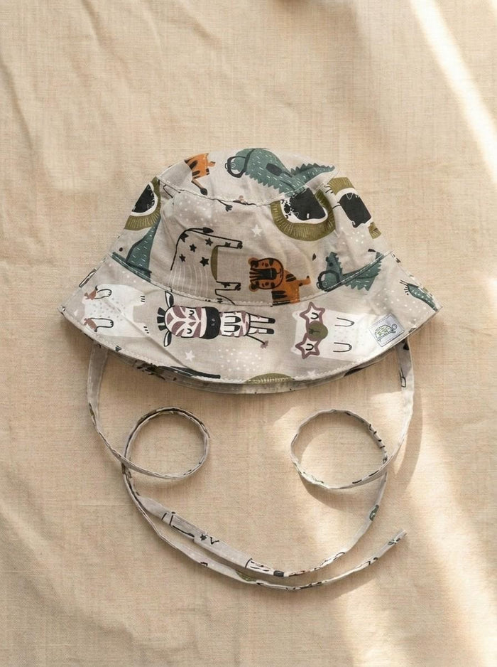 A top-down photograph of a baby bucket hat featuring a light grey or beige fabric covered in a playful zoo animal print, including a zebra, tiger, alligator, and elephant. The hat includes matching fabric chin tie straps and is styled flat on a neutral, textured beige linen background under soft, warm sunlight.