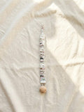 A flat lay photograph of the Zoo pacifier clip, featuring a fabric strap with a white base and a colourful illustration of various zoo animals. The strap has a ruffled, elasticated texture and is attached to a round, natural light-wood clip. It is styled vertically on a neutral, draped cream linen background in soft sunlight.