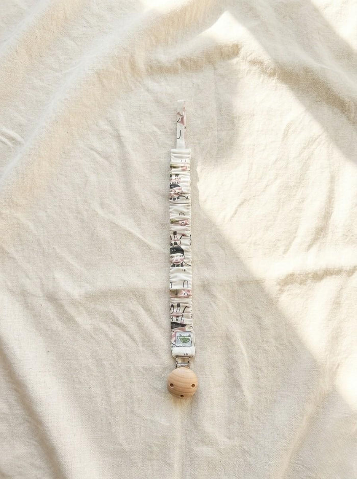 A flat lay photograph of the Zoo pacifier clip, featuring a fabric strap with a white base and a colourful illustration of various zoo animals. The strap has a ruffled, elasticated texture and is attached to a round, natural light-wood clip. It is styled vertically on a neutral, draped cream linen background in soft sunlight.